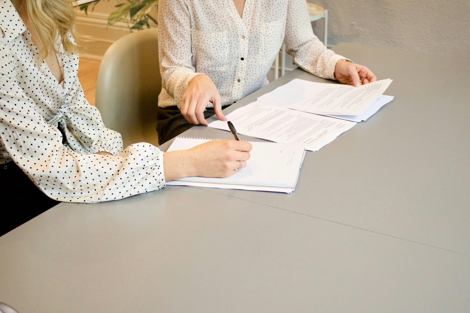 Une femme signe sur une feuille de papier blanc à côté d'une autre femme qui s'apprête à toucher les documents.